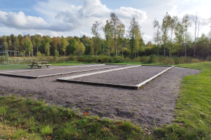 Ein grasbewachsener Platz mit einer Picknick-Tafel in der Mitte, umgeben von Bäumen unter einem klaren blauen Himmel, mit Spielgeräten wie Pfosten und einer Bank auf der linken Seite.