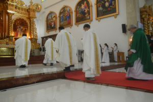 Eine Gruppe von Priestern geht einen roten Teppich in einer Kirche entlang, mit Menschen, die auf dem Boden und auf der rechten Seite stehen, und Foto Rahmen im Hintergrund.