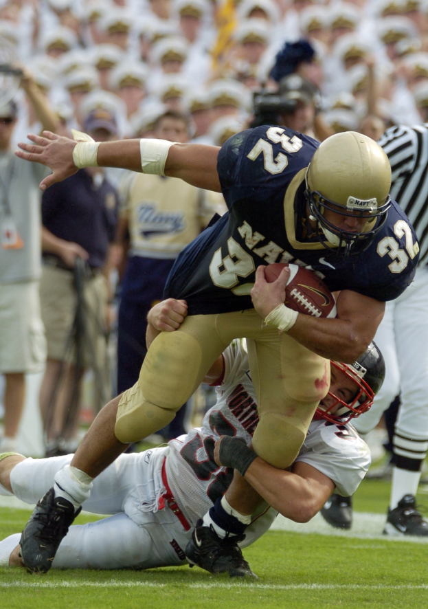 Zwei Fußballspieler auf dem Feld: Einer im Sportdress und Helm mit dem Ball, der von einem anderen Spieler zu Fall gebracht wird, der auf dem Boden liegt, mit anderen Spielern und Zuschauern im Hintergrund.
