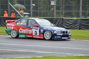 Ein Auto fährt auf einer Rennstrecke mit Text und Zahlen darauf, umgeben von Gras, mit einem Maschendrahtzaun, der mit schwarzem Stoff bedeckt ist, Menschen in orangefarbenen Jacken mit Schirmen und Bäumen im Hintergrund.