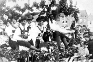 Eine Gruppe von Menschen in traditioneller bayrischer Tracht sitzt auf einem mit Laub geschmückten Festwagen bei einem Umzug, einige halten Musikinstrumente, mit Gebäuden und einem klaren Himmel im Hintergrund, dargestellt in Schwarz-Weiß.