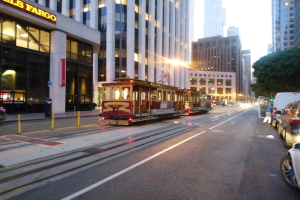 Eine Seilbahn fährt eine Straße in San Francisco entlang mit Fahrzeugen, einem Fußgänger und einem geparkten Van; im Hintergrund sind Gebäude, Bäume und der Himmel zu sehen.