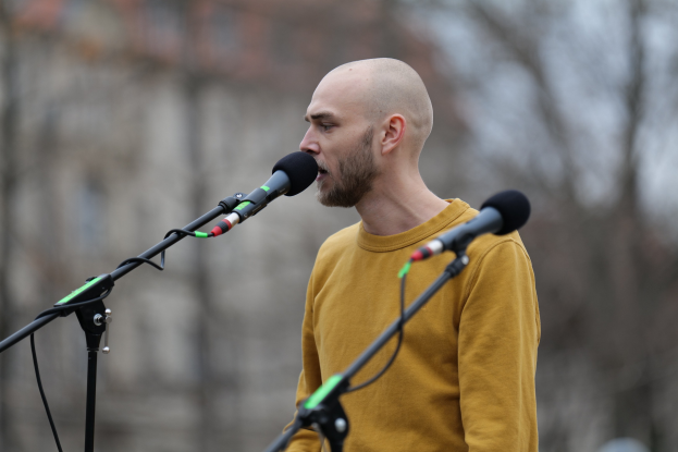 Ein kahlkopfeter Mann in einem gelben T-Shirt singt in ein Mikrofon mit einem unscharfen Hintergrund.