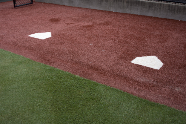 Baseballfeld mit Kunstrasen, umgeben von einem Zaun, mit Home Plate und einer Wand im Hintergrund.
