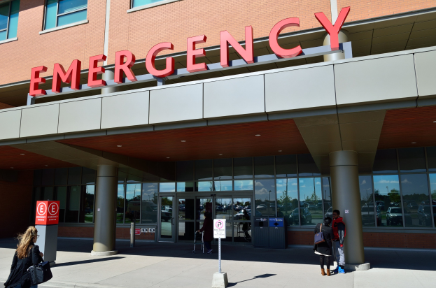 Eine Gruppe von Menschen vor einem Glaswand-Notfallgebäude mit Säulen, einem Namensschild, Schildern auf Pfählen und einem Mülleimer.