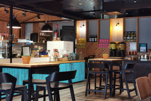 Innenraum eines Restaurants mit Holztischen, Stühlen, einer Topfpflanze auf einem Tisch, einer Schiefertafel, einem Kühlschrank, einem Feuerlöscher, Regalen mit Flaschen, Schränken mit Gegenständen, einem gerahmten Foto an der Wand, Deckenleuchten und Lampen.