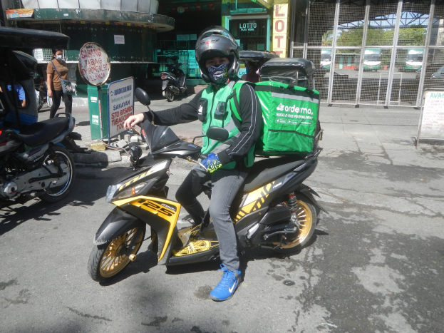 Ein Mann in Helm und Handschuhen sitzt auf einem gelben und schwarzen Roller mit einer grünen Tasche auf dem Rücken, umgeben von Fahrzeugen, Menschen, einer Tafel, einem Mülleimer, einem Gebäude, einem Tor, Bäumen und dem Himmel.