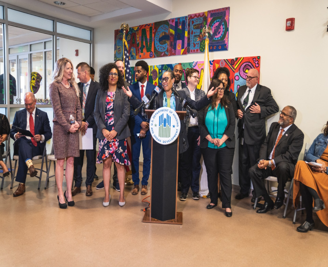 Gruppe von Menschen vor einem Podium mit einem Mikrofon und einer Tafel mit der Aufschrift "Sancho Stadtrat tritt dem Stadtrat bei", mit sitzenden und stehenden Teilnehmern, Fahnen, Feuerloescher und Deckenleuchten im Hintergrund.