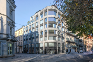 Großes modernes Bürogebäude mit zahlreichen Fenstern, umgeben von Straßeninfrastruktur und Fahrzeugen, mit Fußgängern und Bäumen im Vordergrund unter einem bewölkten Himmel.