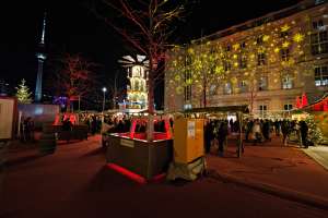 Ein geschäftiger Weihnachtsmarkt in Berlin, Deutschland mit Menschen um geschmückte Stände, Lichter, Tannenbäume, Gebäude, Laternenmasten und einen Turm unter einem dunklen Himmel.