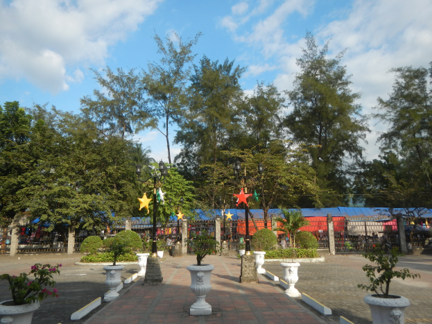 Ein Park mit Topfpflanzen und Bäumen, umgeben von Straßenlaternen, Dekorationselementen, Grills und einer bewölkten Himmel.