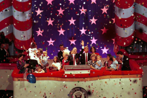 Eine Gruppe von Menschen auf einer Bühne mit einem Rednerpult, das ein Logo, Luftballons und Konfetti zeigt, mit Sternen und einem Vorhang im Hintergrund.