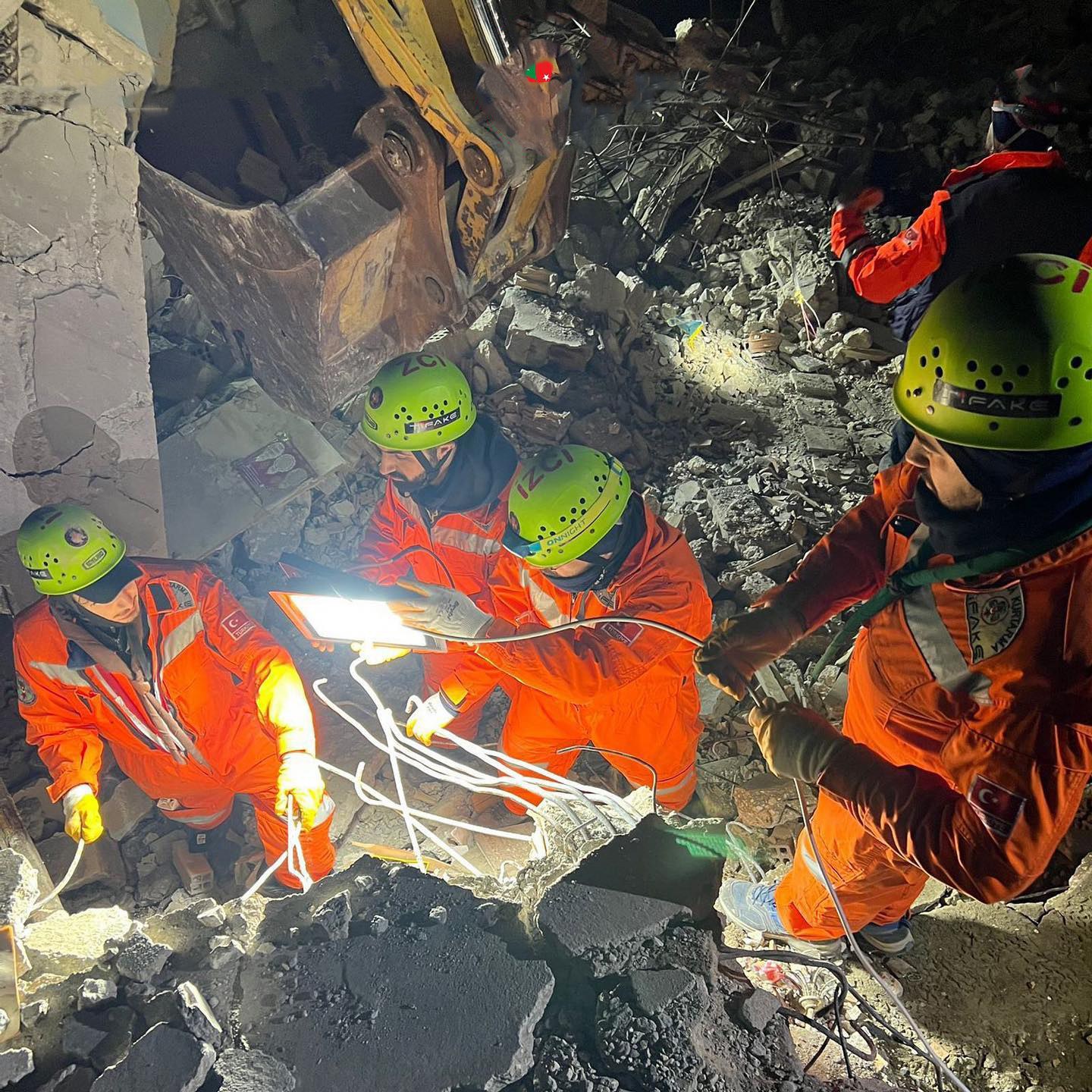 Gruppe von Menschen in orangefarbener Uniform und grünen Helmen bei der Arbeit an Schutt mit einem Kran im Hintergrund während einer Rettungsaktion.