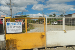 Ein gelbes Tor mit einem Sicherheitshinweisschild, dahinter befinden sich Strommasten, Bäume, Schuppen, Fahrzeuge und ein bewölkter Himmel.