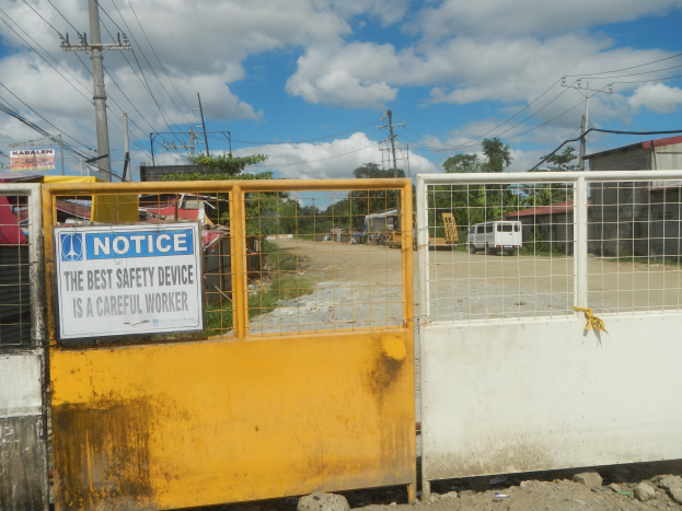Ein gelbes Tor mit einem Sicherheitshinweisschild, dahinter befinden sich Strommasten, Bäume, Schuppen, Fahrzeuge und ein bewölkter Himmel.