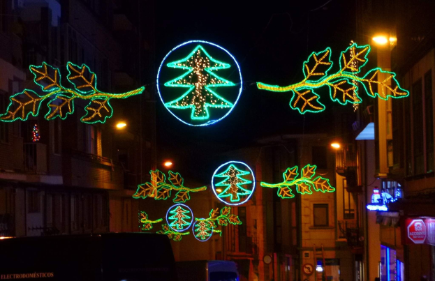 Eine städtische Straße, die mit festlichen Weihnachtslichtern geschmückt ist, die Blätter und andere Dekorationen zeigen, mit Fahrzeugen, die an Gebäuden auf beiden Seiten vorbeifahren.