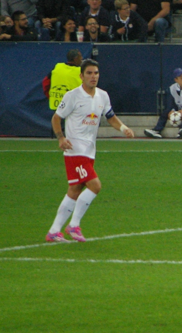Ein professioneller Fußballspieler in weißem T-Shirt und roter Hose, der auf einem Feld mit Zuschauern im Hintergrund spielt.