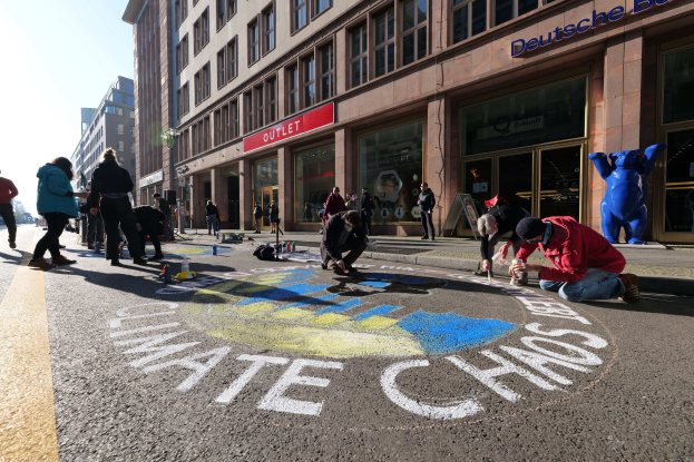 Eine Gruppe von Menschen sitzt vor einem Gebäude mit Fenstern und Namensschildern auf dem Boden, umgeben von Flaschen und anderen Gegenständen, während sie an einer Klimaprotestaktion in Berlin teilnehmen, die von Bäumen und einem klaren blauen Himmel umgeben ist.