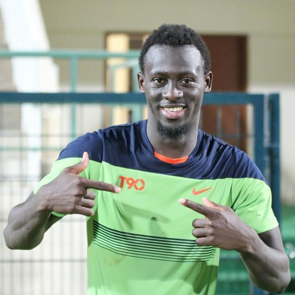 Ein Mann in einem grünen und blauen Hemd lächelt, während er in die Kamera zeigt, mit einem unscharfen Geländer, Wand und Tür im Hintergrund.