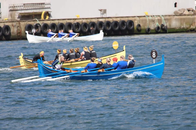 Eine Gruppe von Menschen in einem blauen und gelben Boot, das auf dem Wasser paddelt, mit einer Reifen-linierten Wand und einem Gebäude im Hintergrund.