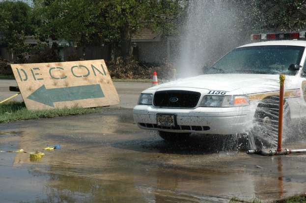 Ein Polizeiwagen fährt durch eine Pfütze, mit einem Schild, Verkehrskegel und anderen Gegenständen am Boden, mit Bäumen, Gebäuden und einem klaren blauen Himmel im Hintergrund.