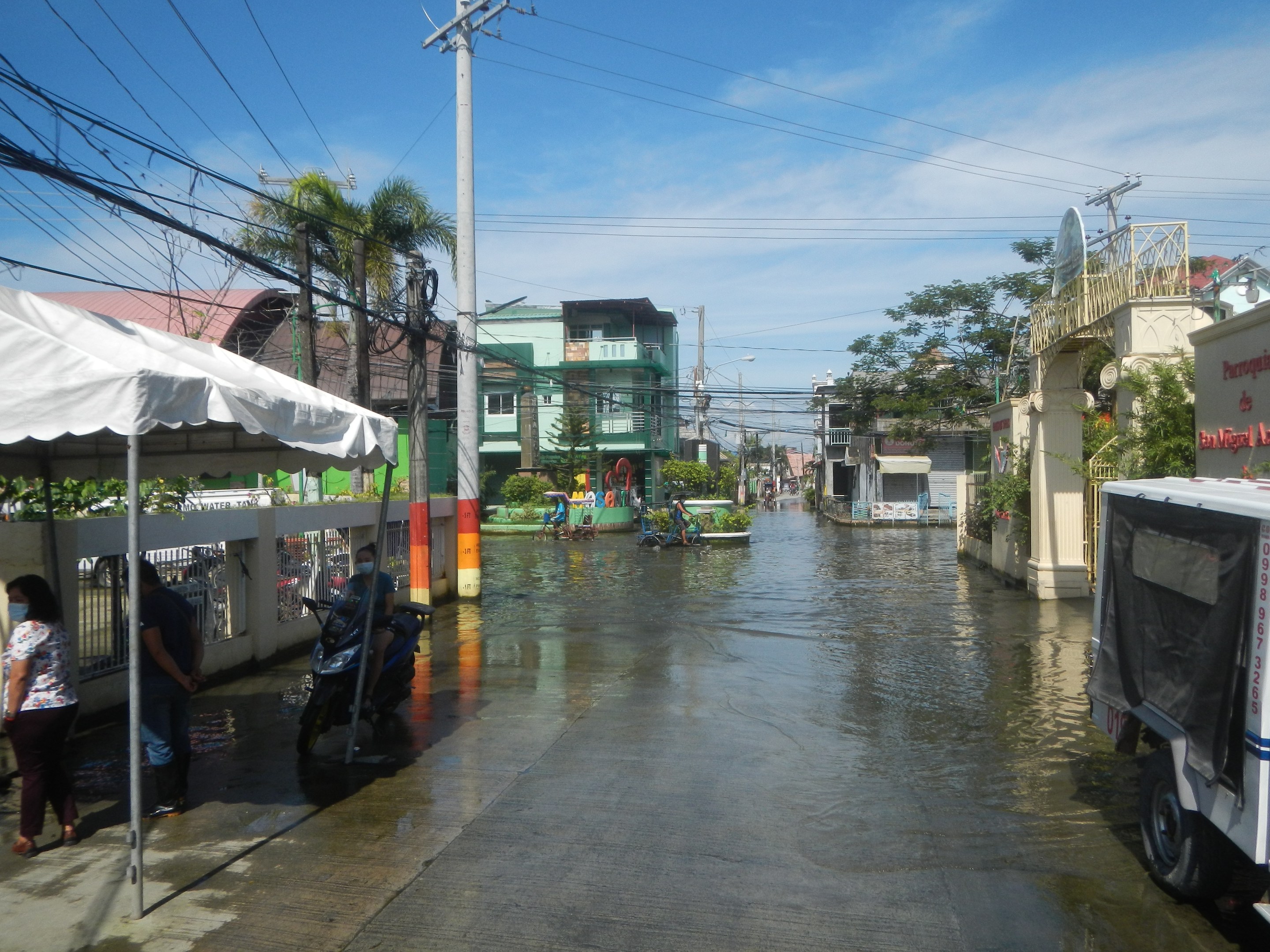 Eine überflutete städtische Straße mit Fahrzeugen, Fußgängern, einem Zelt, Strommasten, Gebäuden, Bäumen und einem bewölkten Himmel.