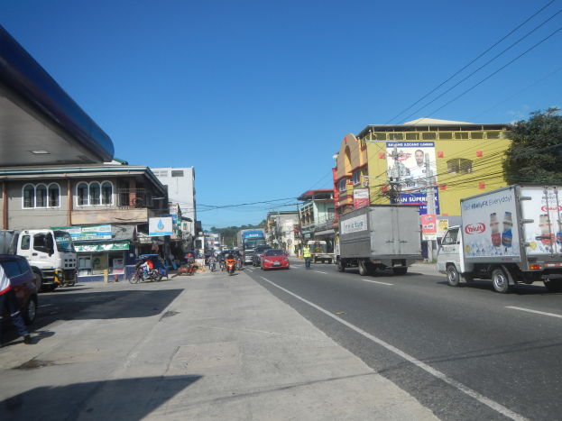 Eine belebte Stadtstraße mit zahlreichen Autos, Lastwagen und Fußgängern auf dem Gehweg, umgeben von Gebäuden, Schildern, Strommasten, Bäumen und einem bewölkten Himmel.