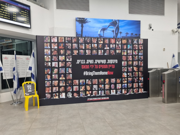 Eine große Wand zeigt verschiedene Bilder, einen gelben Stuhl, israelische Regierungsflaggen auf Stangen, eine Tafel mit dem Text "Bring Them Home Now", einen montierten Bildschirm, Deckenleuchten und andere Gegenstände.
