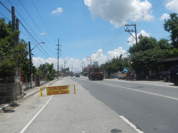 Eine Straße mit Fahrzeugen, ein "Verlangsamen Sie die Schule"-Schild, Strommasten, Bäume, Laternenpfähle und ein bewölkter Himmel im Hintergrund.
