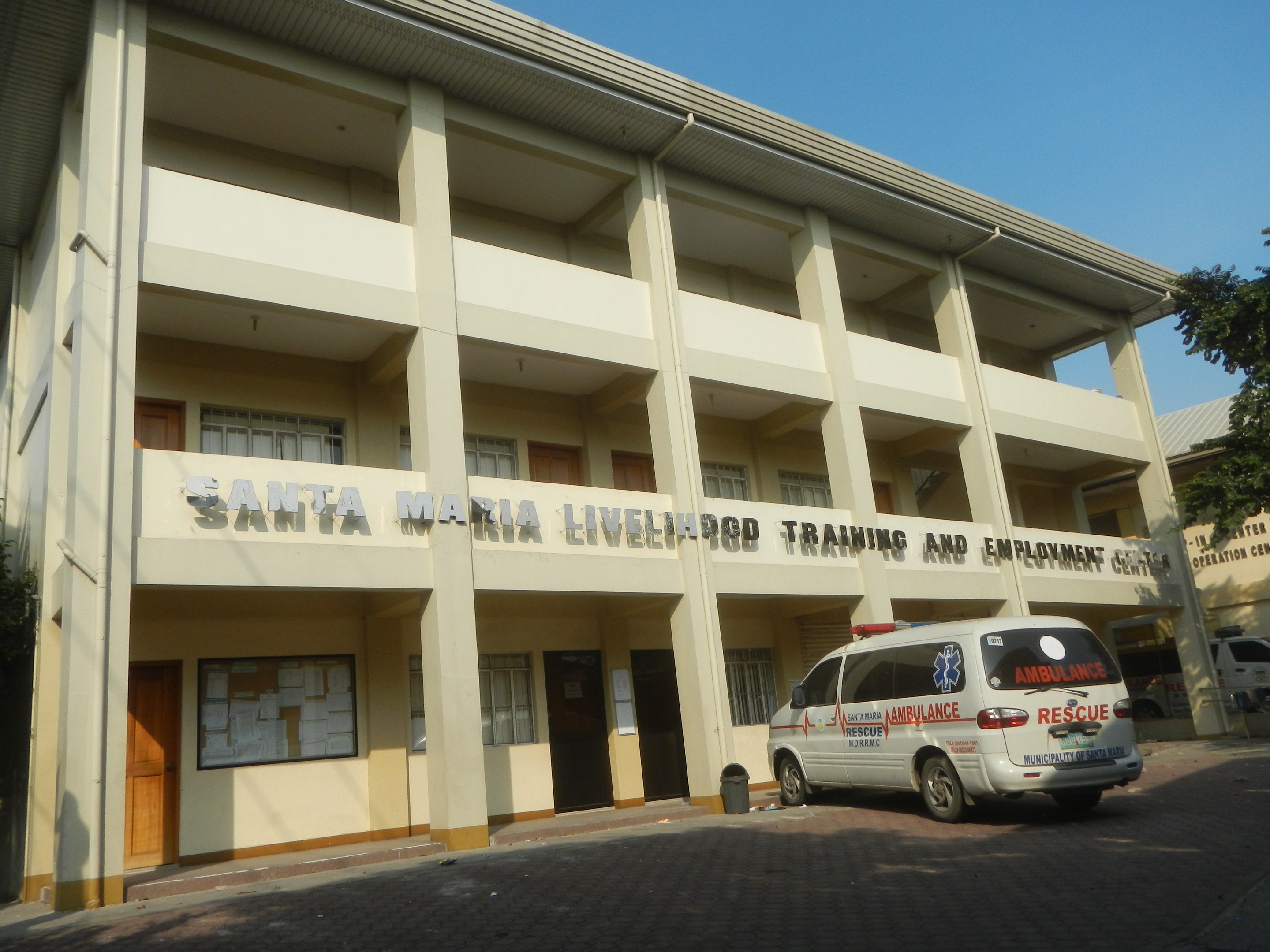 Außenansicht des Santa Maria Lebensmittelausbildungs- und Beschäftigungszentrums mit Fenstern, Türen und Beschriftung, vor einem klaren blauen Himmel mit Bäumen und parkenden Fahrzeugen.