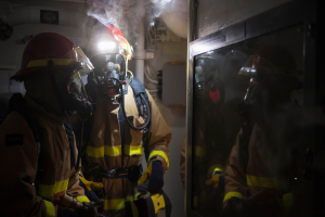 Eine Gruppe von Feuerwehrleuten in Gasmasken und Helmen steht vor einer rauchenden Tür, mit Rohren und anderen Gegenständen im Hintergrund, die auf einen begrenzten Raum hinweisen.