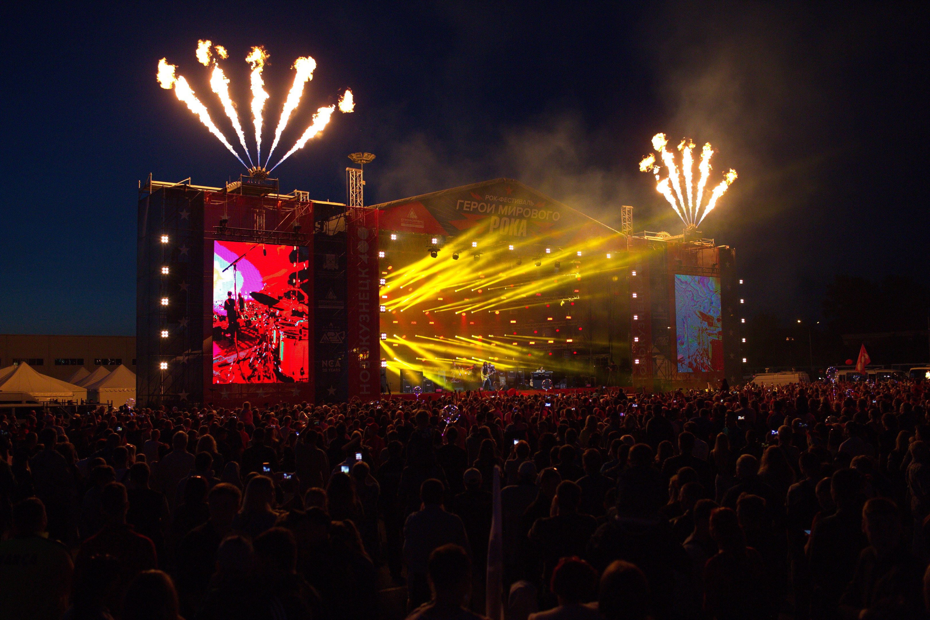 Große Menschenmenge auf einem Musikfestival mit beleuchteter Bühne, Performern, Bannern, Zelten links und Feuerwerk am dunklen Himmel.