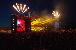 Große Menschenmenge auf einem Musikfestival mit beleuchteter Bühne, Performern, Bannern, Zelten links und Feuerwerk am dunklen Himmel.