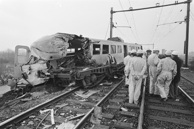 Schwarzes und weißes Bild eines entgleisten Zuges mit zwei kollidierten Zügen auf den Schienen, umgeben von Menschen in Helmen, mit Strommasten, Bäumen, Gebäuden und einem klaren Himmel im Hintergrund.