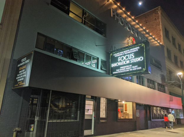 Außenansicht des Focus Innovation Studio in New York City bei Nacht, mit beleuchteten Fenstern, einer beleuchteten Beschriftung, Passanten auf dem Gehweg und Gebäuden im Hintergrund unter einem dunklen Himmel.