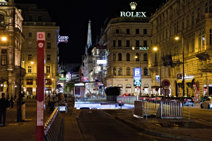 Eine nächtliche Stadtstraße mit fahrenden Autos, Fußgängern auf den Gehwegen, parkenden Fahrrädern, Straßenlaternen, hohen Gebäuden, Absperrungen, Pfosten und einem Rolex-Logo im Hintergrund.
