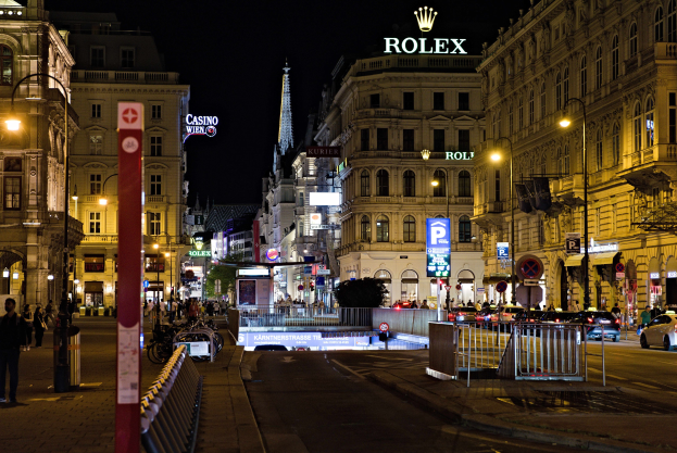 Eine nächtliche Stadtstraße mit fahrenden Autos, Fußgängern auf den Gehwegen, parkenden Fahrrädern, Straßenlaternen, hohen Gebäuden, Absperrungen, Pfosten und einem Rolex-Logo im Hintergrund.