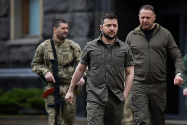 Ein Mann in militärischer Uniform, identifiziert als ehemaliger ukrainischer Präsident Viktor Janukowitsch, wird von zwei bewaffneten Männern in militärischer Uniform vor einem Gebäude mit Pflanzen im Hintergrund eskortiert.