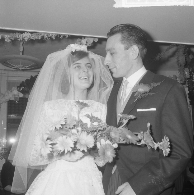 Ein Schwarz-Weiß-Foto eines Brautpaares, das zusammen steht, wobei die Braut einen Strauß hält und der Bräutigam einen Anzug trägt, umgeben von ein paar Menschen und dekorativen Gegenständen im Hintergrund.