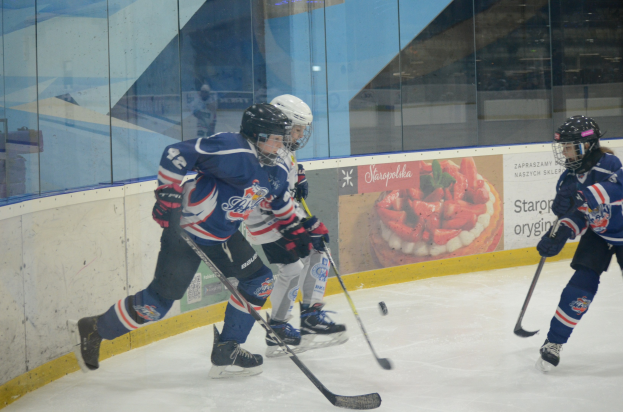 Gruppe junger Menschen, die Eisockey auf einer Indoor-Eisfläche spielen, mit Helmen, Sportbekleidung und Hockeystöcken, sowie einem Plakat im Hintergrund an einer Glaswand.