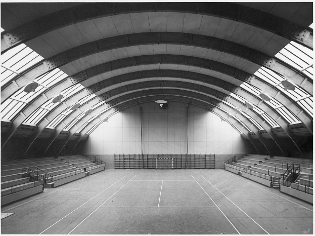 Schwarz-weiß-Foto eines Indoor-Tennisplatzes, der von W├Ąnden umgeben ist, mit Treppen und Gel├Ąndern, die hinaufführen, beleuchtet von Deckenlampen.