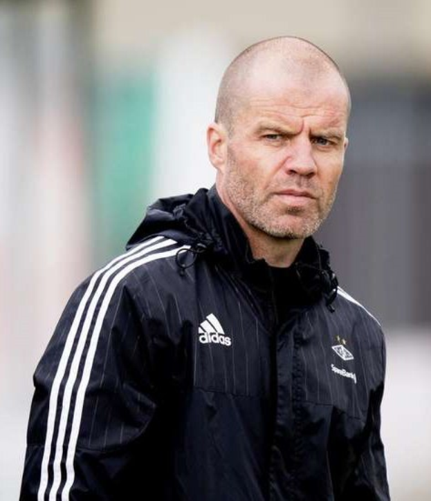 Baldiger Trainer in einer blauen Jacke mit weißen Streifen und weißem Kragen, der auf einem Fußballfeld steht und die Hände verschränkt hält.