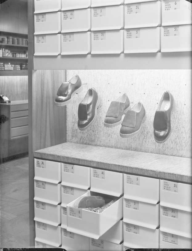 Ein Schwarz-Weiß-Foto eines Schuhgeschäfts mit einer Wandhalterung für Schuhe, Regalen mit verschiedenen Gegenständen links und einer Plattform mit Schuhkartons am Boden.