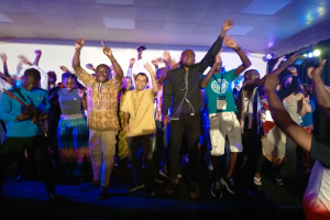 Eine Gruppe von Menschen tanzt auf einer Bühne mit erhobenen Händen, beleuchtet von Scheinwerfern auf der rechten Seite und einem Bildschirm im Hintergrund, unter einer Decke mit Scheinwerfern.