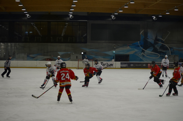 Eine Gruppe von Menschen, die Eis-hockey auf einer Indoor-Eisfläche spielen, alle tragen Helme und halten Hockey-Schläger, mit einer bemalten Wand und beleuchteter Decke im Hintergrund.