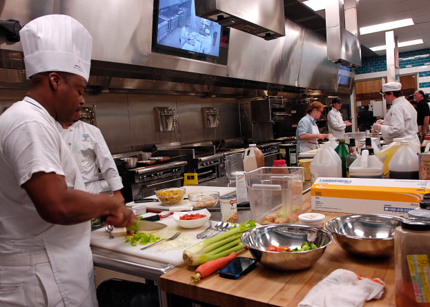 Eine Gruppe von Köchen in einer Küche beim Zubereiten von Speisen auf einem Schneidbrett, mit einem Koch im Vordergrund in einer weißen Schürze und Mütze, der ein Messer hält und von verschiedenen Utensilien, Schüsseln, Gemüse, einem Mobiltelefon, Flaschen und anderen Gegenständen umgeben ist, mit einem Fernseher und Deckenleuchten im Hintergrund.
