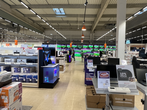 Ein großer Raum in einem Walmart-Geschäft, der mit Elektronik gefüllt ist, die in Regalen angeordnet ist, einschließlich Computer und Drucker, mit Menschen auf dem Boden, einer Säule, Schildern, Deckenleuchten und Deckenventilatoren.