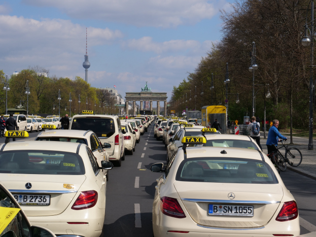 Eine lange Reihe von Taxis, die entlang einer belebten Straße in Berlin, Deutschland, geparkt sind, mit Fahrzeugen, Radfahrern und Fußgängern, flankiert von Laternenmasten und Bäumen, und Gebäuden, einem Bogen und einem Turm im Hintergrund bei einem bewölkten Himmel.