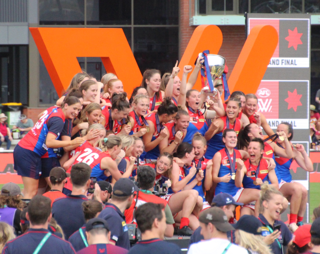 Eine Gruppe von Frauen-AFL-Spielerinnen feiert mit einem Pokal vor einem Publikum, trägt Sportuniformen und hält den Pokal in den Händen. Im Hintergrund befindet sich ein Gebäude mit Glasfenstern und eine Tafel mit Text.