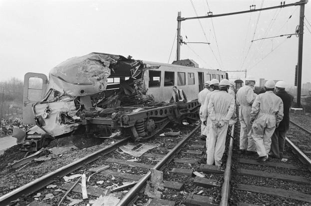 Eine Schwarz-Weiß-Szene eines Zugunfalls mit Menschen in Helmen, die herumstehen, Strommasten, Bäumen, Gebäuden und einem klaren Himmel im Hintergrund.